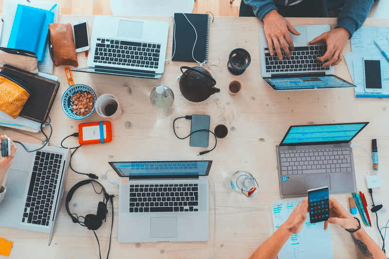 A work table hosts multiple laptops, cell phones, and external hard drives while two people sit and work.