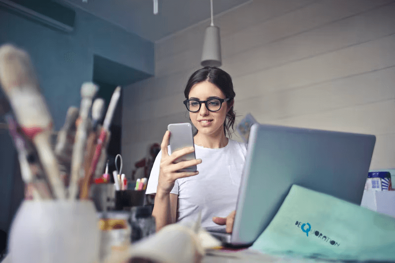 A young woman sits at her laptop while holding her mobile phone to donate using nonprofit online fundraising.