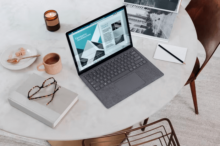 A laptop is open on a round table top, with a notebook and glasses resting off to the side.