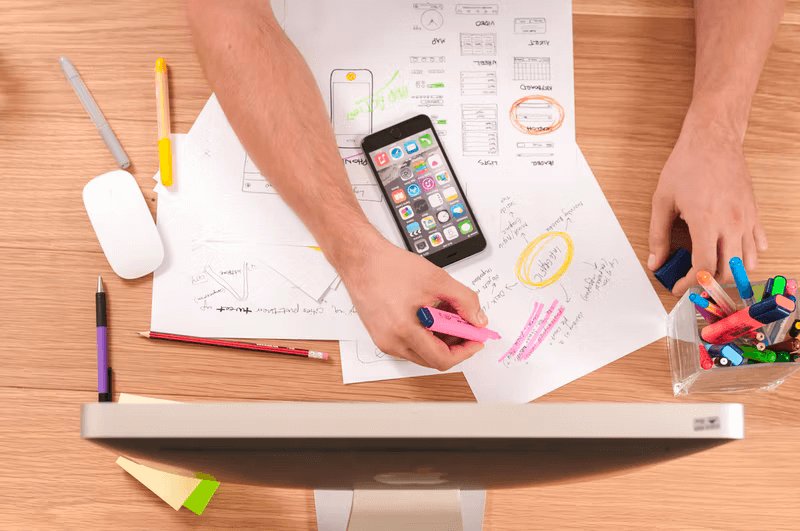 Someone sits at a table with a computer, a mobile phone, research papers, and pens.