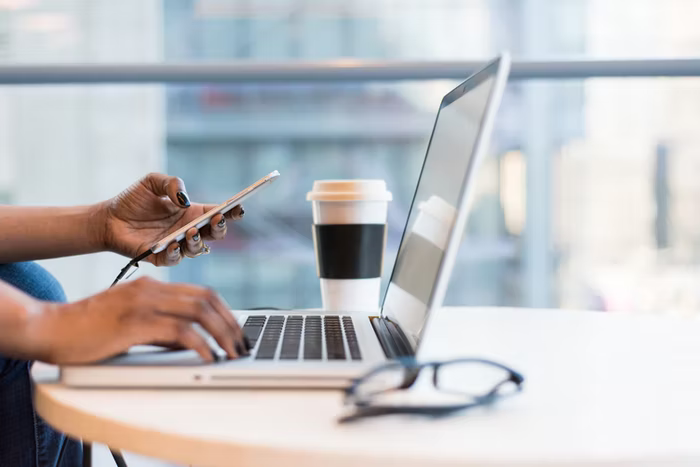 A young woman holds a cell phone in one hand and types on her laptop keyboard with the other.