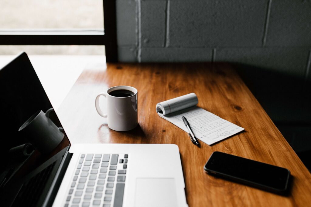 A computer is open on a woodtop desk with a coffee mug and pad and pencil next to it