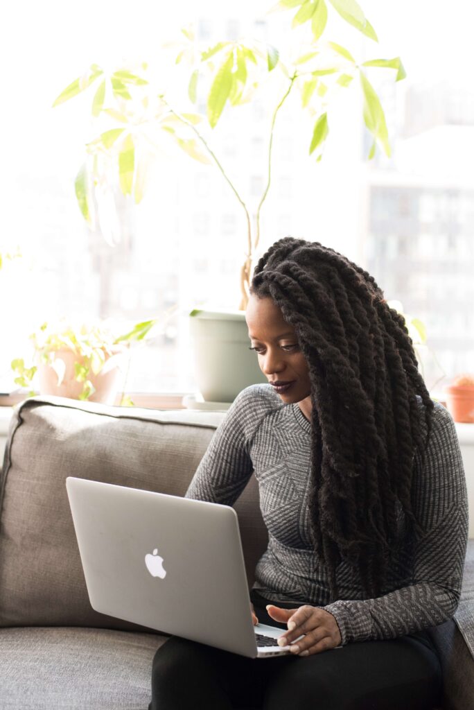 A young Black woman sits on a gray couch as she works on her laptop