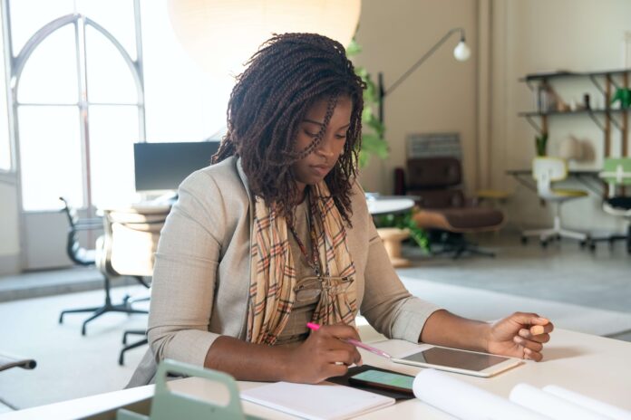 A young Black woman, who is a nonprofit professional, sits at a desk with her smartphone and tablet to work on the organization’s marketing strategy